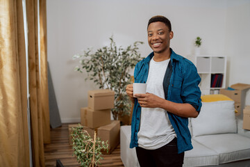 Smiling brunette with dark complexion is standing at window of new apartment, looking out at the view from it, drinking coffee in a mug, living room piled with cardboard boxes with things after move