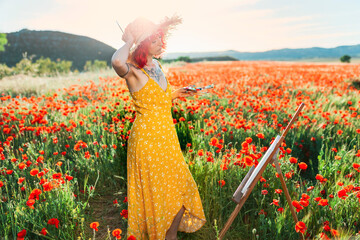 Female artist wearing straw hat looking at canvas on poppy field during sunny day