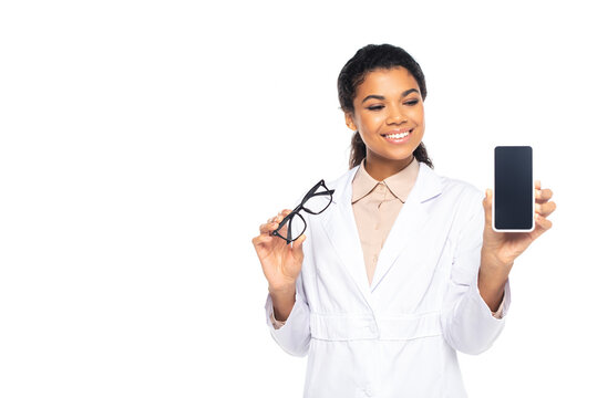 Positive african american ophthalmologist holding cellphone with blank screen and eyeglasses isolated on white.