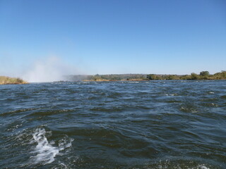 Speed boat cruising on River Zambesi above the mighty Victoria Falls with the waterfall's spray in...