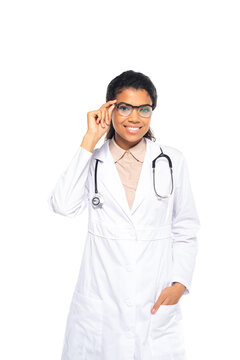 Young african american ophthalmologist holding eyeglasses and hand in pocket of white coat isolated on white.