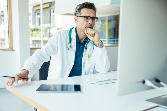Male doctor with hand on chin using computer at office - Powered by Adobe