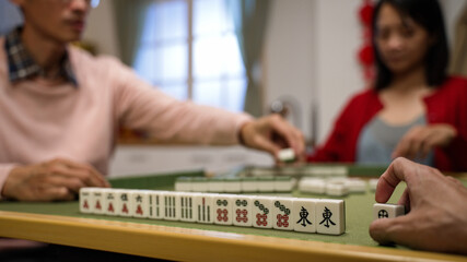 closeup with selective focus of a player's hand playing with a tile next to his hand of mahjong pieces while his family are having their turn. chinese words translation: east