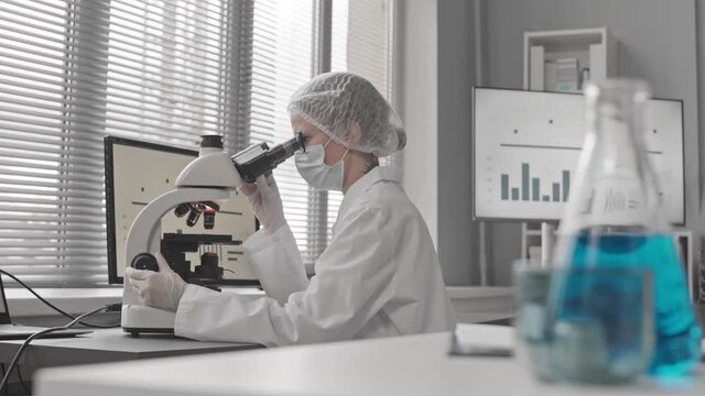 Racking focus from beakers with liquids to young female Caucasian scientist wearing medical uniform and eyeglasses, sitting at desk in laboratory, using microscope, typing on computer