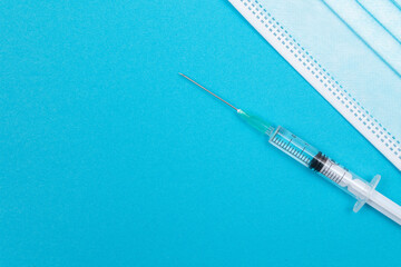 A Syringe and Face Mask on Blue Table - Top View. Syringe is Prepared for Vaccination or Injection. Medical Experiment. Blue Disposable Medical Mask. Virus protection - Flat Lay