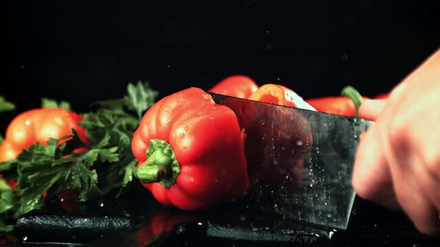 A man's hand cuts a sweet pepper into two halves with a knife. On a black background. Filmed is slow motion 1000 frames per second.