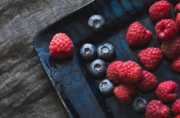 Raspberries and blueberries. Fresh juicy appetizing colorful summer fruits on a navy blue plate, linen tablecloth. Close up photo, background
