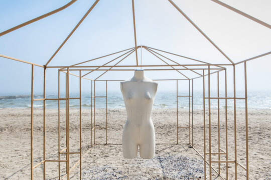 The white headless body of a mannequin hangs in a metal gazebo on the seashore in sunny weather 