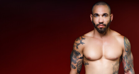 Man of 30-40 years old with beautiful muscular body with tattoos stands on red dark background and looks directly into camera. Advertising banner with space for copying. Studio portrait. © SerPak