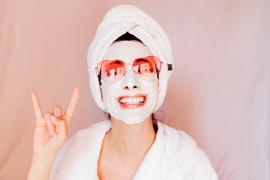 Happy young woman with beauty face mask showing horn sign against peach background