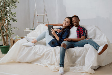 Happy smiling in love young students renting a new apartment, redecorating the room, relaxing on the sofa secured with foil, attractive brunette lies on sofa leaning against the dark-skinned boy