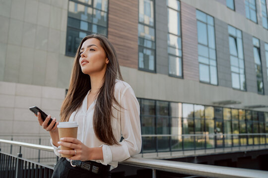 Attractive girl working in an office, corporation, company, dressed in elegant clothes stands leaning against the railing in front of glass building, coffee break, waiting for phone call, looks ahead