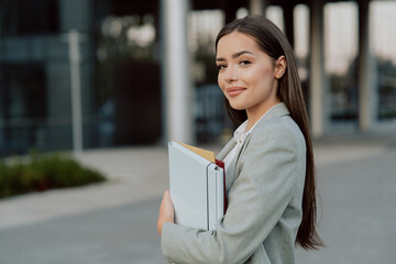 A smiling, pretty, successful girl running her own company manager boss is standing next to glass modern building going to meeting conference to work holding files with documents in her hand