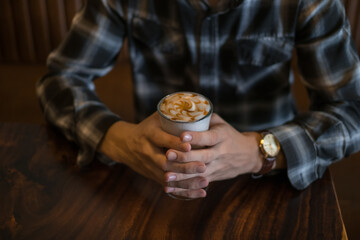 Man in checkered shirt drinks coffee in a bar. Holding caramel latte in his hands. Close up, Coffee shop, cafe, latte, caramel cappuccino.