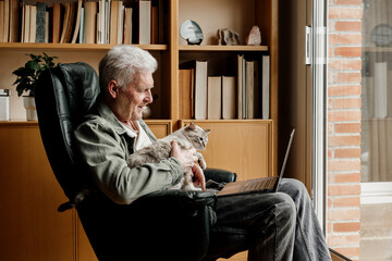 Senior man with laptop holding cat while sitting on chair at home