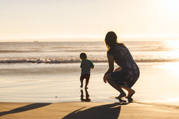 Woman Crouching Behind Son Running On Shore - obrazy, fototapety, plakaty