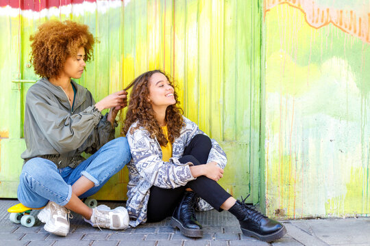 Young woman tying hair of female friend while sitting on skateboard