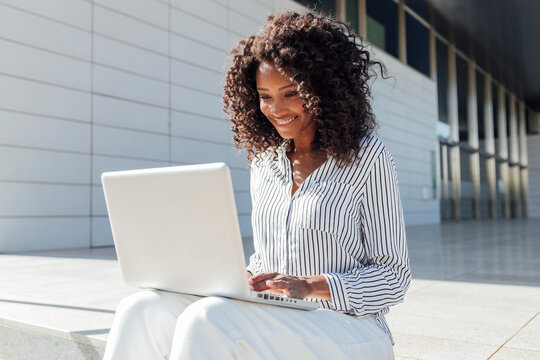 Smiling young businesswoman using laptop while sitting outside office building