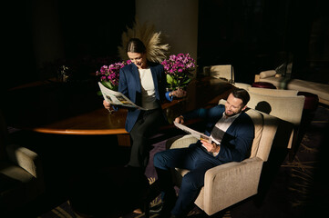 Overhead view of two handsome people, middle aged Caucasian man and beautiful elegant woman in business suit, colleagues, business partners reading a newspaper in the hotel lounge area