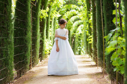 Girl in white communion dress looking down while standing in garden