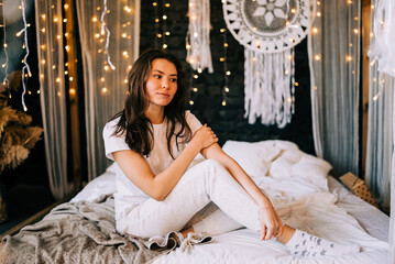 Fototapeta premium Romantic beautiful woman sitting on bed and touching her chin with hand. Portrait of dreamy cute young woman on the bed