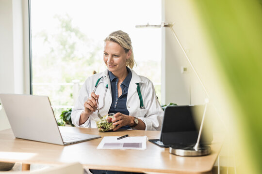 Smiling female doctor eating food while looking at laptop in office