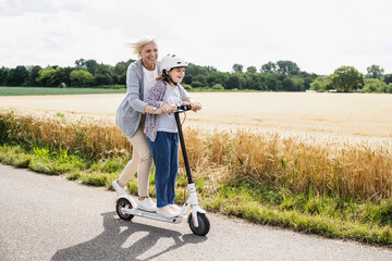 Happy granddaughter and grandmother playing while riding push scooter during sunny day