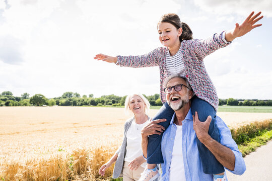 Happy man carrying granddaughter on shoulder while walking on road during sunny day