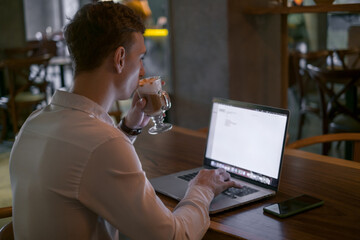 Young caucasian business man surfing the net on laptop while drinking coffee in a bar. Handsome man working in cafe with laptop. Typing on keyboard