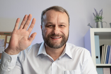 Middle aged man gestures hello at the camera. Make online calls and live video conference chat...