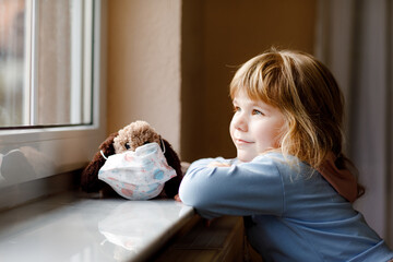 Cute toddler girl sitting by window and looking out. Dreaming child put a medical mask on soft dog toy feeling happy. Lockdown and isolation concept during corona virus pandemic time. Lonely kid. - obrazy, fototapety, plakaty