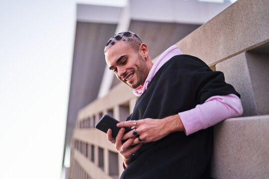 Smiling eccentric male leaning on wall and browsing smartphone