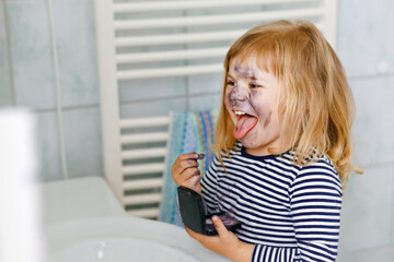 Funny little toddler girl using mother's make up and painting face with eye shadows. Happy baby...