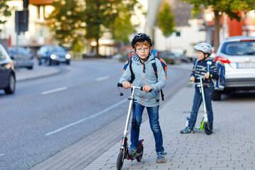 Two school kid boys in safety helmet riding with scooter in the city with backpack on sunny day. Happy children in colorful clothes biking on way to school.
