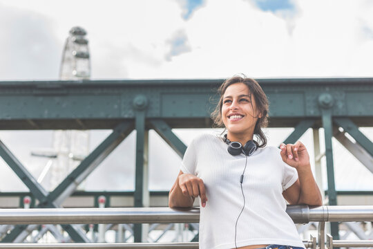 Smiling woman with headphones looking away while leaning on railing at bridge, London, England, UK