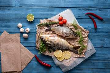 Top view two carp fish are lying on paper on a wooden blue background, next to ingredients, tomatoes, lemons and spices