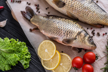 two carp fish are lying on paper on a wooden black background of boards, next to ingredients, tomatoes, lemons and spices