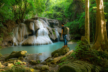 Fototapeta premium Young man with backpack standing near a waterfall in forest. Male hiker in the nature during rain.