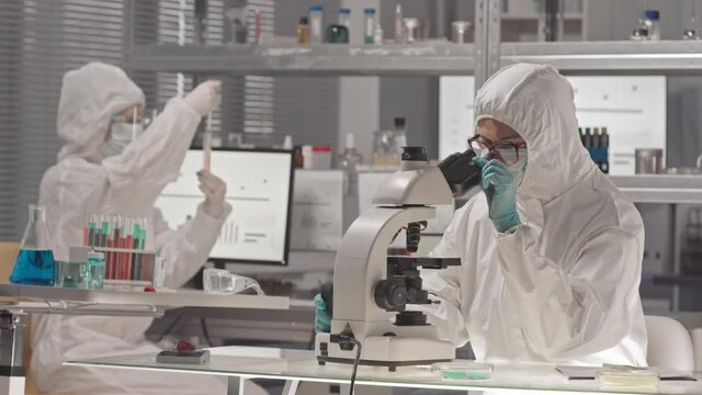 Waist-up of scientist wearing hazmat overall, eyeglasses and mask, sitting at desk in research center, using microscope