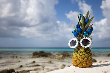 Pineapple with sunglasses decorated with Christmas balls stands on the beach.