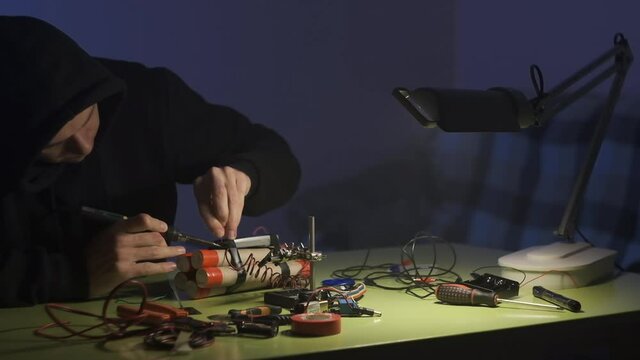 man in black sweatshirt in dark room is soldering and assembling a bomb of dynamite with telephone as detonator