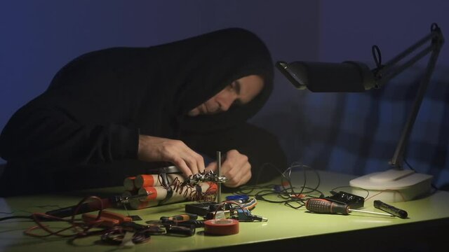 man in black sweatshirt in dark room is soldering and assembling a bomb of dynamite with telephone as detonator