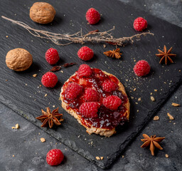 A delicious crouton with jam and whole raspberries on top and some walnuts near it, on a black slate background