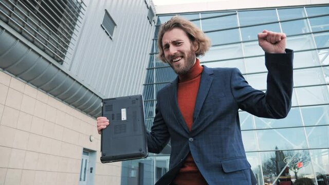 Witty guy dancing celebrating new career going to work with laptop outdoors. Comic playful office manager dancing feeling happy about new startup project walking near business center.