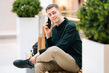 Portrait of confident young man talking on mobile phone sitting on bench in shopping mall looking...