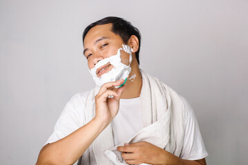 Handsome asian man shaving. Handsome asian man shaving his face and looking at camera while standing on grey background