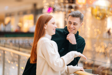 Laughing cute redhead young woman telling to handsome man interest story standing by railing in shopping mall with bright interior. Happy couple spending time together at store during holiday sales.