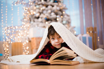 A boy in pajamas reads a book under a Christmas tree . New Year's mood. Reading books . Children's...