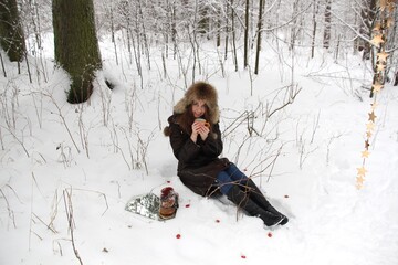 Young woman in brown fur coat and hat sitting in snow frost, holding cup of coffee and smiling in winter forest