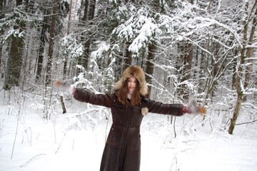 Young attractive woman in brown fur coat, hat, mittens standing with waving hands in snow frost and smiling in russian winter forest